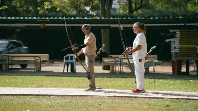 Two mature archers practicing their aim with longbows at an outdoor shooting range. Focused elderly men pulling back their bowstrings