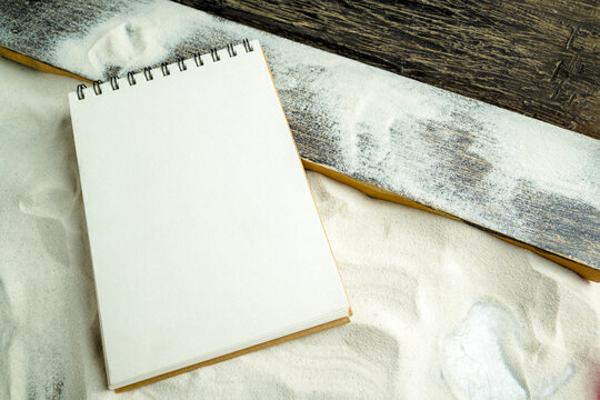 Blank spiral notebook on white sand next to a rustic wooden board, providing copy space for summer travel plans or journaling. Mockup or copy space for travel or vacation