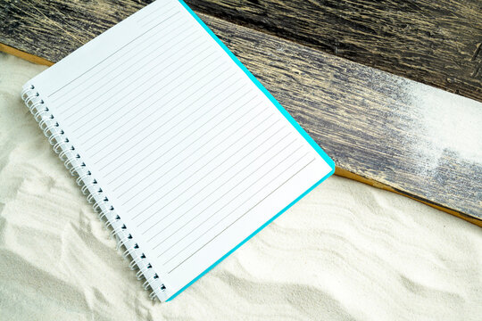Blank lined spiral notebook on white sand next to a rustic wooden plank, ideal for summer planning or travel journaling. Mockup or copy space for travel or vacation