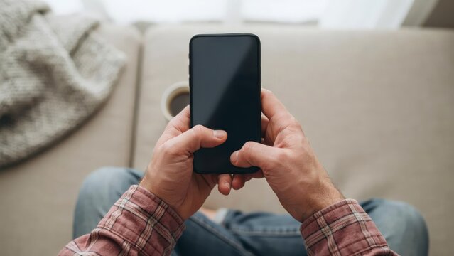 Top view of a man holding a black smartphone with a blank screen while sitting on a couch