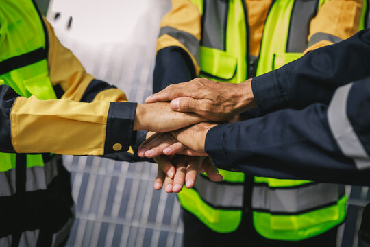 Close-up of construction workers stacking hands together while wearing high-visibility vests and protective work wear, teamwork and workplace safety.