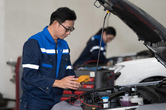Asian male mechanic inspects and repairs battery pack, car engine and components with hood open inside professional service garage