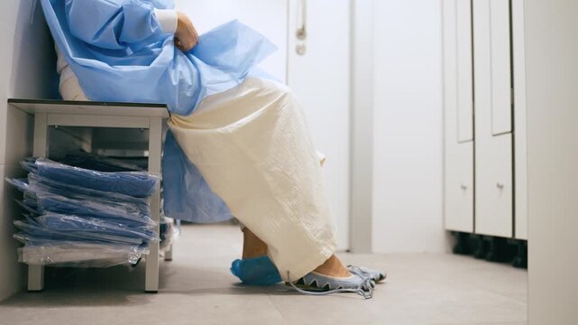 Female doctor sits in a hospital changing room putting on disposable protective shoe covers before entering the operating room