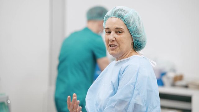 Middle aged woman in a sterile gown and cap sitting in an operating room, talking and smiling while waiting for medical procedure