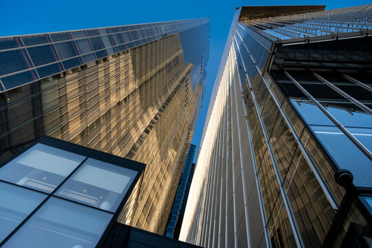 Upward view between modern towers in London within the United Kingdom with reflective glass facades and vertical lines against blue sky emphasizing urban height