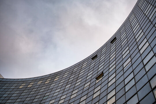 Curved modern glass facade with repeating windows and geometric lines rising beneath a cloudy sky in a dramatic low angle urban architecture composition