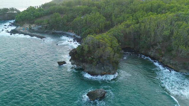 Aerial view of lush green cliffs meeting the turquoise sea with crashing waves along the rugged coastline in Pacitan, East Java, Indonesia.