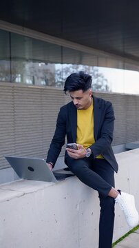 Stylish young Moroccan businessman sitting outdoors on a concrete ledge, working on his laptop while simultaneously using his smartphone. Vertical