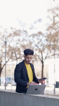 Young Moroccan businessman with a mustache multitasking outdoors. He is using his smartphone and laptop computer while working in the city street. Vertical