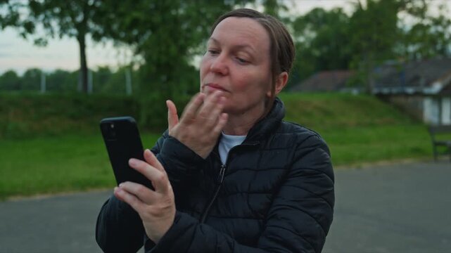 Middle-aged woman outdoors uses her smartphone as a mirror, touching her face and focusing on the screen while standing on a quiet road near grass and trees at dusk. 4K.