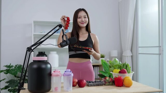 Smiling Asian fitness influencer recording a health podcast, scooping pink protein powder into a shaker bottle. Woman vlogger creating sports nutrition and healthy diet content with fresh food.