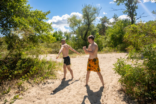Two Young Athletic Men in Swim Shorts Warming Up and Exercising on a Sandy Beach on a Sunny Summer Day