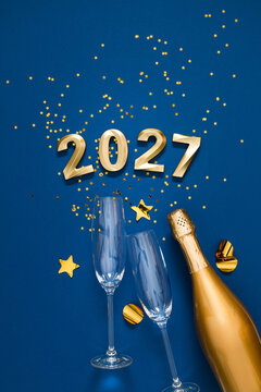 Happy New Year 2027 vertical celebration flat lay with golden numbers, champagne bottle, flutes, and star confetti ornaments on deep blue background with elegant copy space for holiday text.