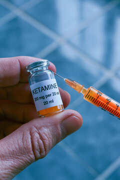 Person's hand holding a small glass vial and a syringe filled with an orange liquid. The vial is labeled "ketamine" in black letters.