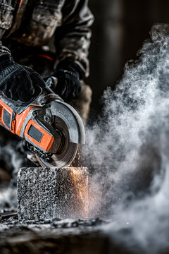 A worker in dark, soiled work and black gloves uses a handheld orange and grey circular concrete saw.