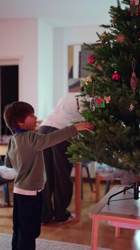 Young boy hangs an ornament on the Christmas tree in the living room, helping decorate the family home while celebrating tradition, anticipation and festive joy