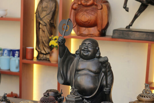 Close-up of a bronze Laughing Buddha statue holding a fan and a ruyi scepter on a blurred souvenir shop shelf background.