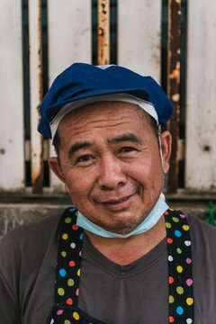 Smiling mature Asian man wearing a blue chef hat and colorful polka dot apron with a face mask pulled down