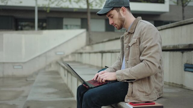 Focused young man typing on a laptop sitting on concrete steps outdoors in the city