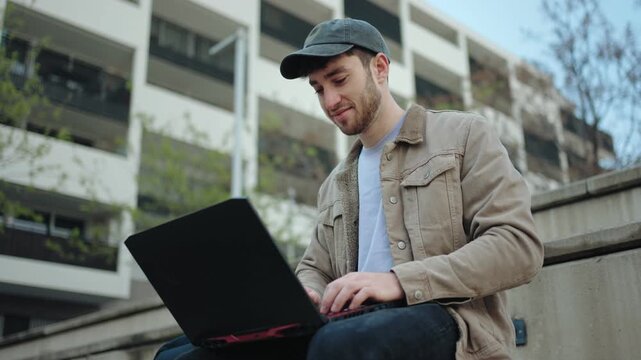 Casual man using laptop computer sitting outdoors working in the city