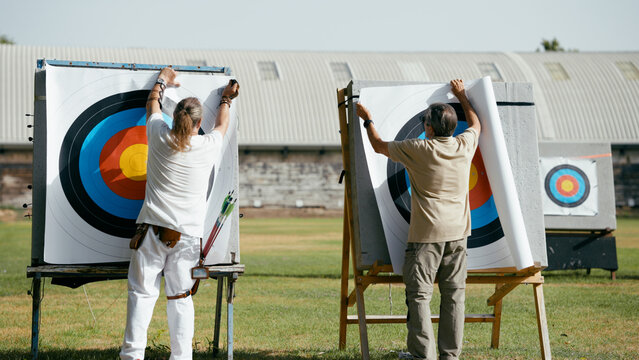 Two mature men replacing paper targets on archery stands outdoors, preparing for practice and focusing on their aiming skills