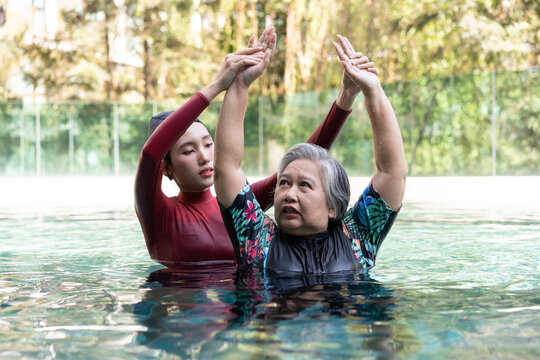 Senior woman practicing aquatic therapy with a young female instructor in a swimming pool, raising both arms for guided mobility exercise while promoting healthy aging and daily wellness.