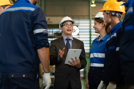 A factory supervisor speaks with industrial workers wearing protective helmets inside a manufacturing facility. The scene highlights workplace communication, leadership, safety, and management.
