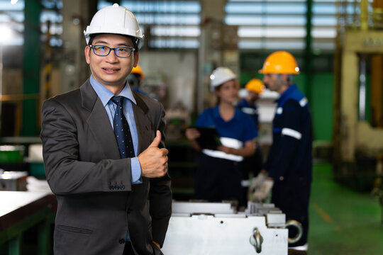 A smiling factory engineer wearing a hard hat gives a thumbs-up inside a manufacturing facility while workers appear in the background. Concept of workplace confidence, teamwork,  industrial success