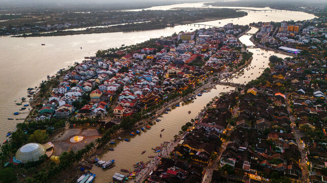 Aerial view of the historic town of Hoi An, featuring traditional yellow buildings, tiled rooftops, and the Thu Bon River with boats at sunset in Hoi An, Da Nang, Vietnam.