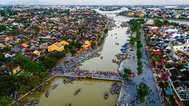 Aerial view of Hoi An Ancient Town with traditional wooden boats on the Thu Bon River, historic yellow buildings, and people walking along the waterfront in Hoi An, Da Nang, Vietnam.