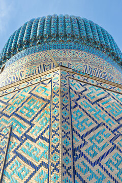 Gur-e-Amir mausoleum dome and tilework detail in Samarkand, Uzbekistan. Ribbed turquoise dome with intricate geometric mosaic patterns on brick walls under clear blue sky. Ideal for travel.