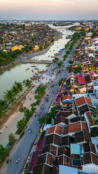 Aerial view of the historic town of Hoi An, featuring traditional yellow buildings, the Thu Bon River with small boats, and people walking along the riverside in Hoi An, Da Nang, Vietnam.