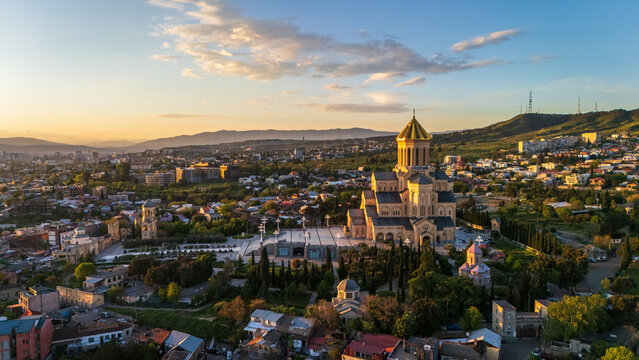Aerial view of the Holy Trinity Cathedral of Tbilisi, also known as Sameba, surrounded by urban landscape, greenery, and distant mountains under a golden sky in Tbilisi, Tbilisi, Georgia.