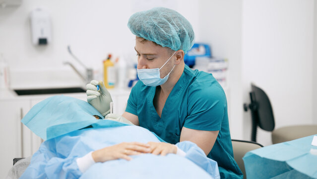 Surgeon wearing scrubs, mask, and cap, performing eye surgery on a patient covered with drapes in a medical setting