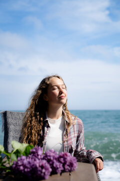 Young woman relaxing by the sea with closed eyes, enjoying fresh air and sunlight. Casual seaside lifestyle scene with lilac flowers on the table, representing relaxation and appreciation of nature.