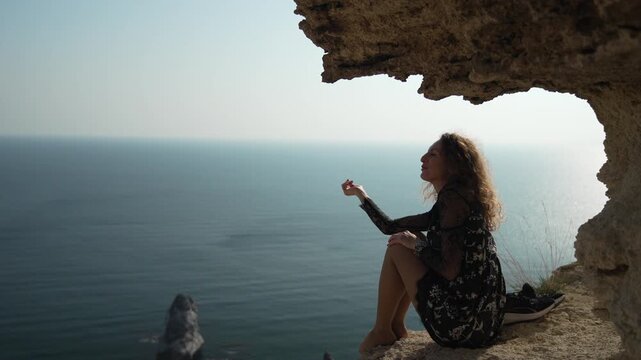 Woman cliff ocean, enjoying freedom while sitting on high rock ledge above the sea, laughing happily.