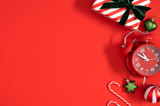 Christmas flat lay with red alarm clock, striped gift box, candy canes, and festive bauble ornaments on vivid red background with negative space for text for holiday marketing and banners.