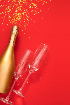 Vertical New Year flat lay with golden champagne bottle, empty flutes, and star confetti on vibrant red background with elegant copy space for festive holiday greetings and design layout.