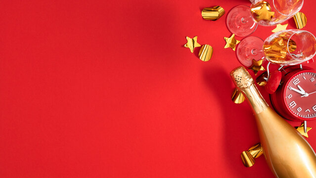 New Year Eve party flat lay banner with golden champagne bottle, flutes, red alarm clock, and star confetti on vibrant red background with large creative copy space on left side for festive text.