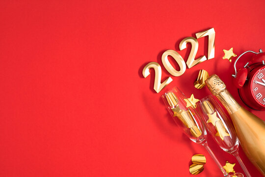 Happy New Year 2027 celebration flat lay with golden numbers, champagne bottle, flutes, alarm clock, stars and confetti ornaments on vibrant red background with copy space for text