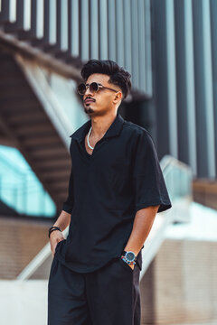 Moroccan man posing with hands in pockets, wearing trendy black streetwear, round sunglasses, and a pearl necklace in an urban setting