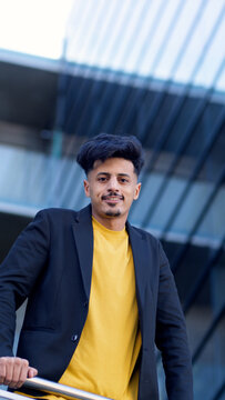 Young Moroccan man with dark hair and a beard, wearing a black jacket over a yellow shirt, smiling in an urban environment