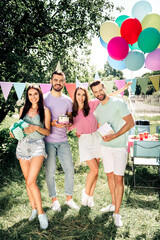 Naklejka premium Friends celebrating a birthday outdoors with balloons, cake, gifts and bunting at a sunny summer garden party — happy young adults enjoying a relaxed picnic celebration