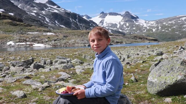 Child, eating lunch before starting the hiking of Kyrkja mountain on a beautiful sunny summer day, sunset, snow, lakes, valley