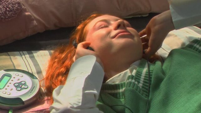 Handheld shot of Caucasian teenage girl lying on bed next to school notebooks, putting on headphones connected to portable CD player. Ideal for retro tech and youth lifestyle campaigns