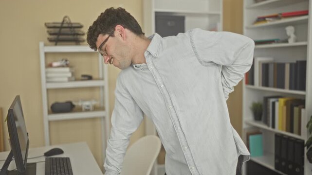 Man holding lower back with one hand while leaning on desk beside a monitor in an office building, showing backache; work stress.