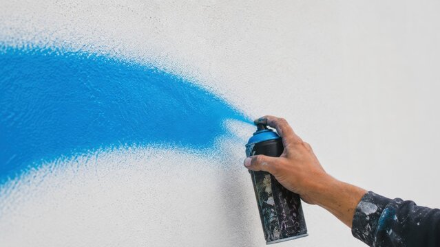A person's hand uses a blue spray paint can to create a stroke on a textured white wall, representing street art or graffiti.