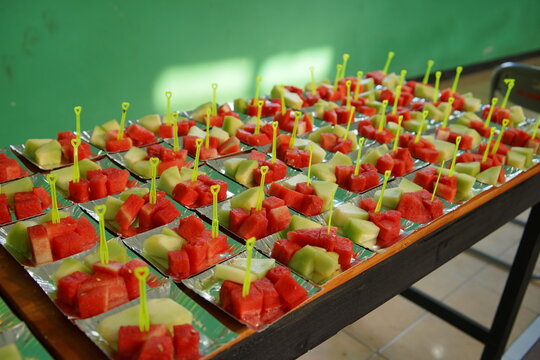 Row of small plates with sliced fresh watermelon and melon pieces served with small yellow plastic forks, healthy fruit snack buffet setup for events or school gatherings