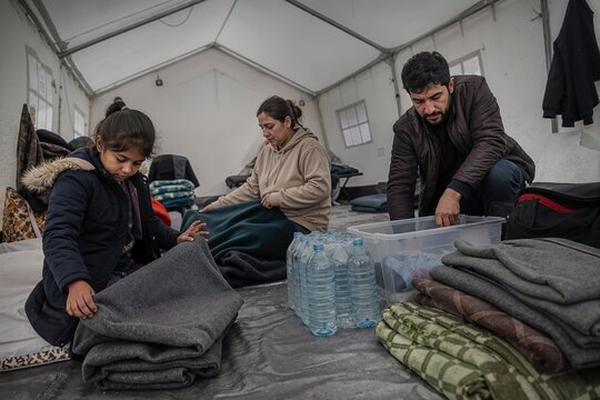 Family organizing blankets and water inside a temporary disaster relief shelter. Displaced people arranging supplies in an emergency tent. Humanitarian aid and survival concept