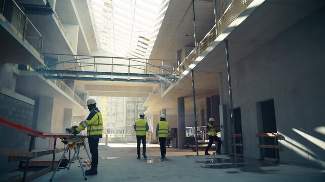 Wide View of Construction Site Interior Where Two Engineers Walk Away Using a Digital Tablet Computer While a Builder Operates a Saw and Crews Continue Work on Commercial Real Estate Development